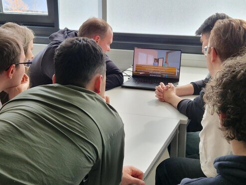 Studierende sitzen um einen Laptop und sehen sich eine Szene des Spiels an