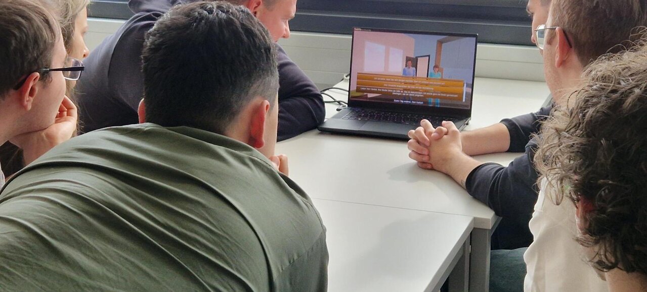 Studierende sitzen um einen Laptop und sehen sich eine Szene des Spiels an